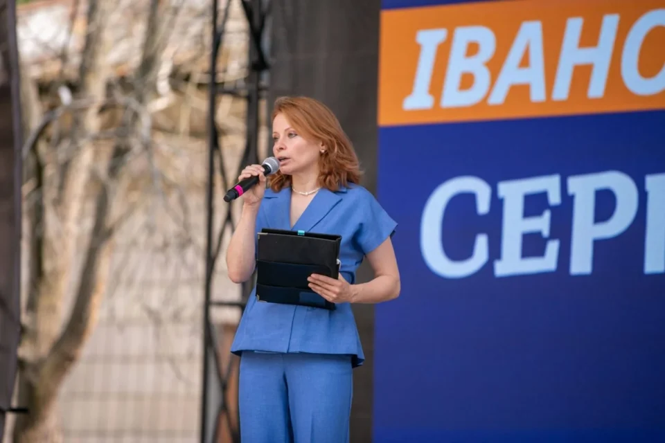 Жінка в блакитному костюмі говорить у мікрофон біля банера Івано-Франківська