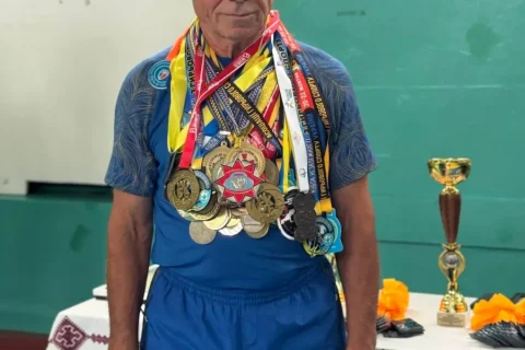 Пожилой мужчина в синем спортивном костюме с множеством медалей на груди на фоне зелёной стены в спортзале