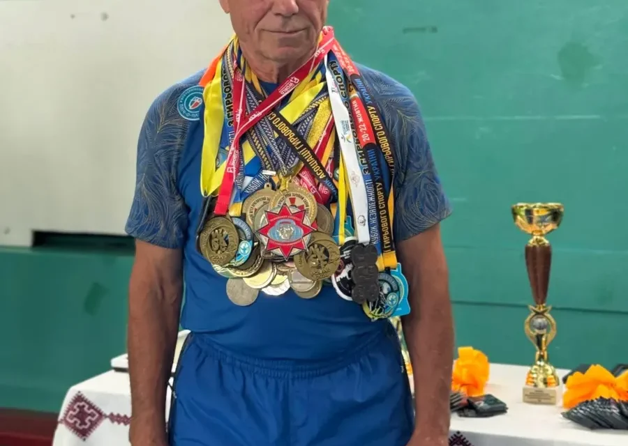 Пожилой мужчина в синем спортивном костюме с множеством медалей на груди на фоне зелёной стены в спортзале